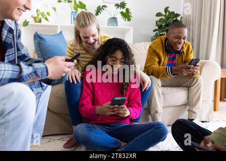 Happy diverse group of teenage friends sitting on couch using smartphones at home Stock Photo