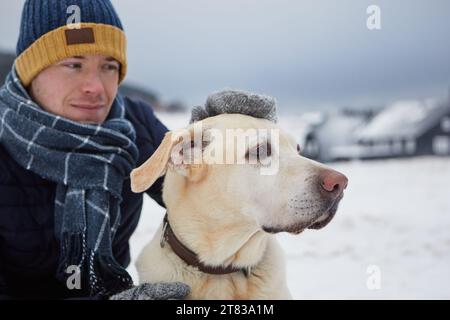 Man dressed in warm jacket, with scarf and cap stroking his dog. Pet owner and labrador retriever against mountain village in winter. Selective focus Stock Photo
