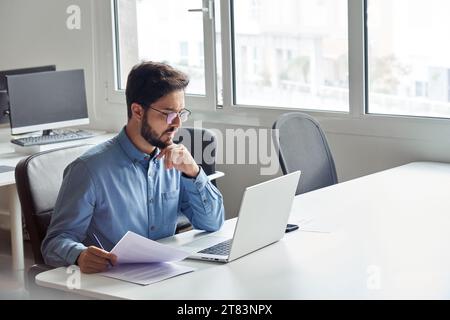 Busy young business man working on laptop computer holding documents. Stock Photo