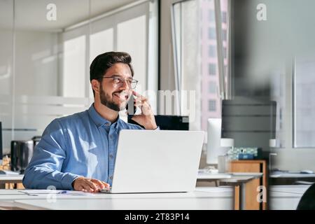 Young happy busy latin businessman talking on cell phone using laptop in office. Stock Photo