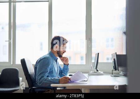 Busy serious young business man working on laptop computer holding documents. Stock Photo