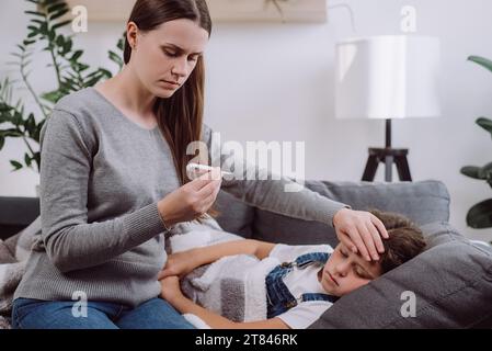 Worried young caucasian mother treating sick daughter kid suffering from flu and fever, holding thermometer, checking high body temperature, applying Stock Photo