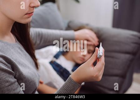 Close up of caring young mother treating sick little daughter kid suffering from flu and fever lying on couch, holding thermometer, checking high body Stock Photo