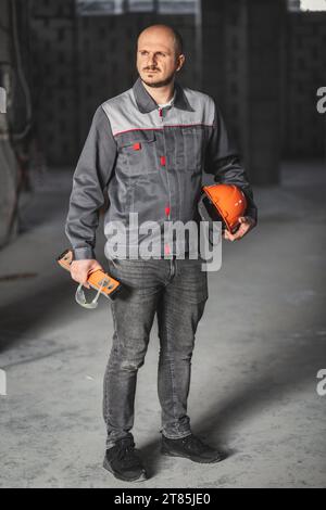Construction worker in overalls, with a helmet, a construction level and safety glasses in his hands, is ready for his shift. Professional at work Stock Photo
