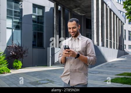A young successful male businessman in a shirt walks outside an office building, holds a phone in his hands, uses an application on a smartphone, reads online pages, smiles happily Stock Photo