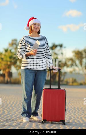 Independent mature woman in Christmas Santa Claus hat, striped shirt holds passport, boarding ticket, suitcase on seashore with palms backdrop celebra Stock Photo