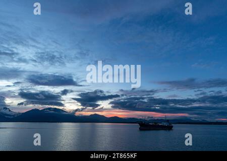 Cargo tanker sits anchored in beautiful remote port surrounded by calm waters as sun sets in pink hues behind mountains Stock Photo