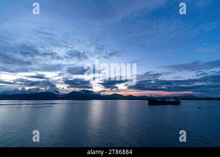 Cargo tanker sits anchored in beautiful remote port surrounded by calm waters as sun sets in pink hues behind mountains Stock Photo