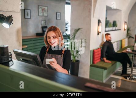 Beautifull smiling waitress dressed black uniform processing customer orders using point of sale order terminal system touch screen. Bartender working Stock Photo