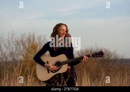 beautiful young smiling woman playing an acoustic guitar while standing in field among tall dried grass. Talented musician. Autumn sunset. Romantic mo Stock Photo