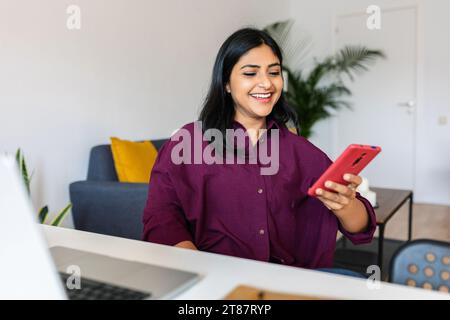 Happy indian woman using mobile phone sitting with laptop on table at home Stock Photo
