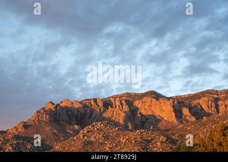 The Sandia Mountains illuminated by the light of the setting sun, casting the rocks in orange and pink colors, with a light sky with high, thin clouds Stock Photo