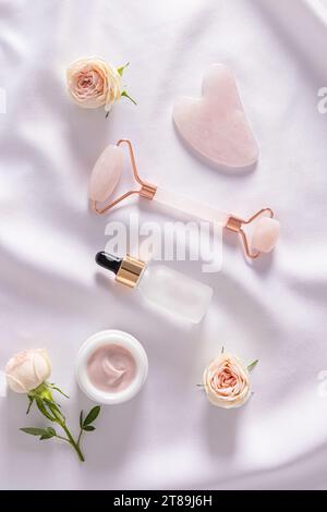 Rose quartz roller massager, gua sha scraper, cosmetic glass matte bottle on delicate white satin. rose flower. Beauty concept. . Top Vertical view Stock Photo