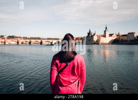 Female traveler dressed in purple coat standing near the Vltava river in Prague (Czech Republic) and looking on Charles Bridge on sunset - photo from Stock Photo