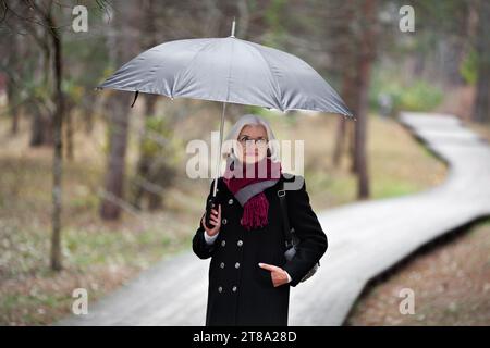 Attractive beautiful middle aged woman with umbrella stands in autumn park. Mature female walks along path in forest, dressed in stylish clothes black Stock Photo