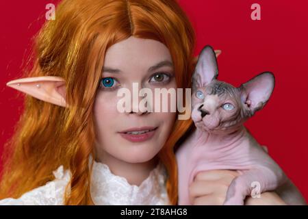 Portrait of ginger young woman cosplay elf in white dress with expressive eyes of different colors, holding Sphinx kitten. Woman with long curly hair Stock Photo