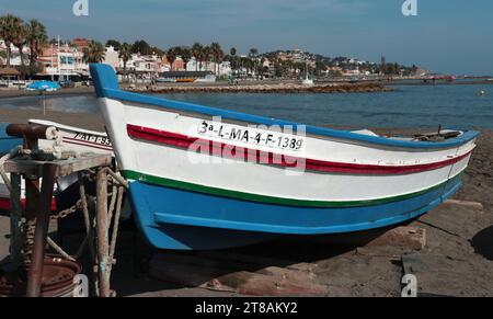 Pedregalejo fishing village, Malaga, Andalucia: traditional painted wooden rowing boat for fishing sits on the sand; seafront promenade behind Stock Photo