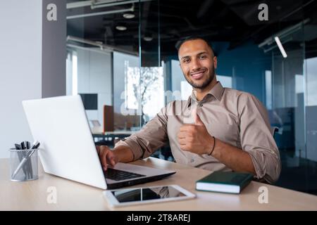 Happy african american businessman sitting in light office at desk, working on laptop, showing thumb up, smiling and looking at camera. Stock Photo