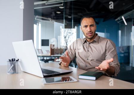 Dissatisfied hispanic male office worker sitting at desk indoors at desk, working on laptop, spreading hands surprised indignant, looking at camera. Stock Photo