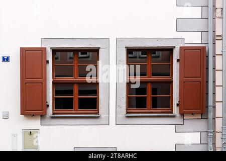 Two rectangular windows with brown frames and wooden shutters against a white wall with a drainpipe. From the Windows of the world series. Stock Photo