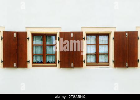 Two rectangular windows with brown frames and wooden shutters on a white wall background. From the Windows of the world series. Stock Photo