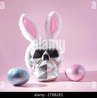 Skulls in Springtime: A Metallic Silver Skull Head Wearing Pink Easter Bunny Ears and Glittery, Sparkling Easter Eggs on a Pastel Pink Background Stock Photo