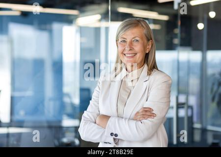 Portrait of mature successful senior woman boss inside office in business suit, businesswoman smiling and looking at camera, standing by window with crossed arms, banker finance woman. Stock Photo