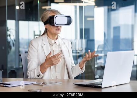 Senior experienced business woman using virtual reality glasses for remote meeting, video call, female boss serious talking with colleagues in virtual world, working throughout office with laptop. Stock Photo