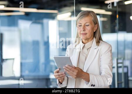 Serious senior woman using tablet computer, businesswoman in business suit senior experienced thinker standing near window, inside office, boss financial investor. Stock Photo
