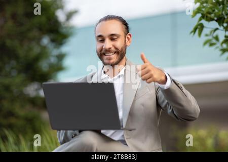 Young Businessman Using Laptop Outdoors, Making Video Call And Showing Thumb Up Stock Photo