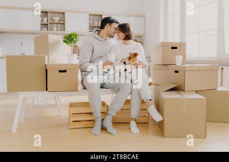 Affectionate couple with a dog enjoying a coffee break amid moving boxes in a bright kitchen Stock Photo