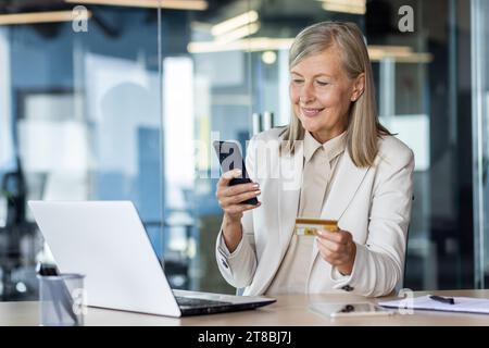 Senior businesswoman working in office at desk, using mobile phone and credit card. Stock Photo