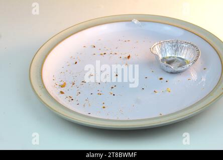 A photo of an empty silver mince pie tin on a white plate covered in crumbs on a white desk. Stock Photo