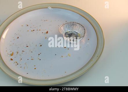 A photo of an empty silver mince pie tin on a white plate covered in crumbs on a white desk. Stock Photo