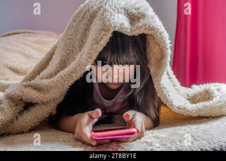 A child using a smartphone lying in bed, playing games, watching online videos, scrolling the screen. Children's screen addiction. Children's room. Stock Photo