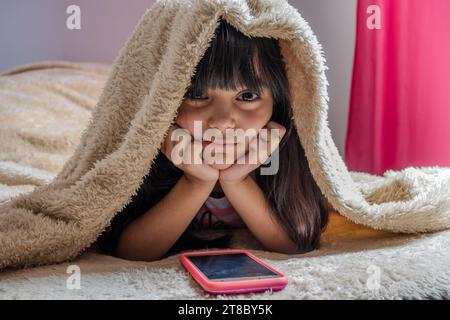 A child using a smartphone lying in bed, playing games, watching online videos, scrolling the screen. Children's screen addiction. Children's room. Stock Photo
