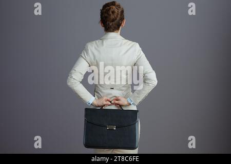 Seen from behind small business owner woman in a light business suit with briefcase against gray background. Stock Photo