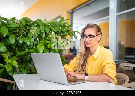 Focused woman with laptop sitting in cafe near green plant. Female freelancer works on laptop sitting at table in cafe. Modern woman working on laptop sitting in cafe. Modern lifestyle Stock Photo