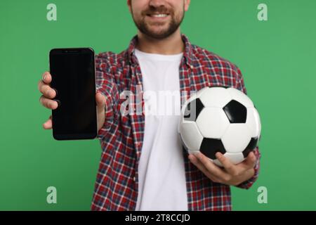 Sports fan with ball and smartphone on green background, closeup Stock Photo
