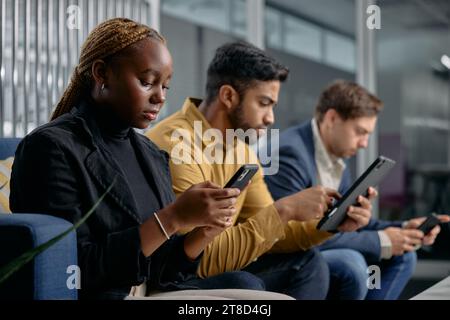 Three multiracial young adults in businesswear sitting on sofa and using technology in office Stock Photo