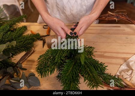 Top view composition of female hands making Christmas wreath from natural branches of spruce, pine, eucalyptus. Girl wraps green twigs onto Kraft base Stock Photo