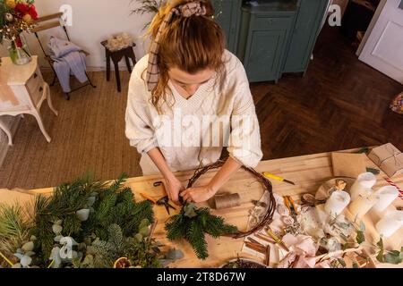 Top view of young woman making handmade Christmas wreath at home from natural materials. DIY home decoration for New Year holidays. Blogger writes DIY Stock Photo
