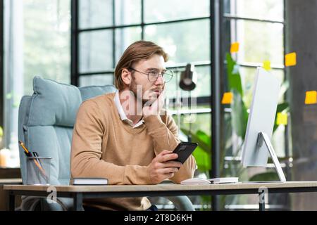 Upset man sitting at the table with a phone in his hands looks tiredly at the computer screen, holds his head with his hand, overworked burnout. Stock Photo