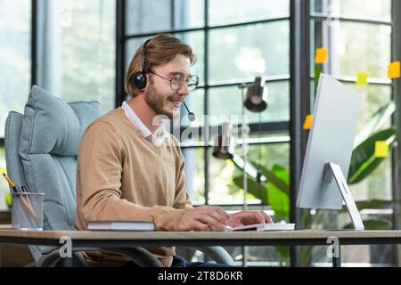 Handsome male programmer sits in a headset in the office at the table in profile, works at the computer, smiles and looks at the screen. Stock Photo