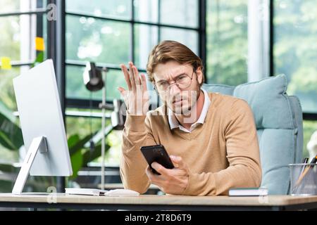 Angry businessman holding phone, shouting and waving hand, sitting at desk in office, upset about bad news, angry at non-working phone. Stock Photo
