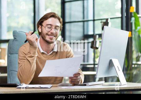 Satisfied young guy doing paperwork in the office, sitting at a desk in front of a computer, joyfully going through business papers, smiling Stock Photo