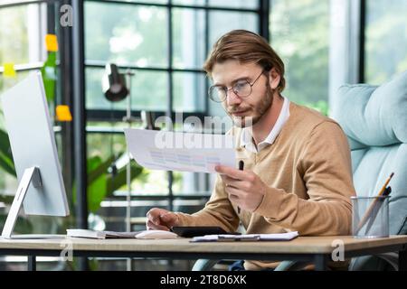 A young man is concentrating on work schedules, sitting at a desk in a bright office and doing paper work, using a calculator while concentrating on work. Stock Photo