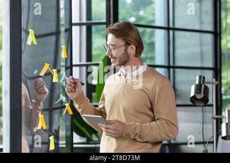 Happy man in casual clothes standing near a glass note board and sticking colorful stickers with reminders and plans on it, writing something with a pen, office work planning concept. Stock Photo