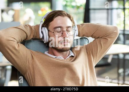 Portrait of a tired office worker in headphones resting at the workplace in a chair, arms crossed on the title with closed eyes, napping during a break between work, listening to music. Stock Photo