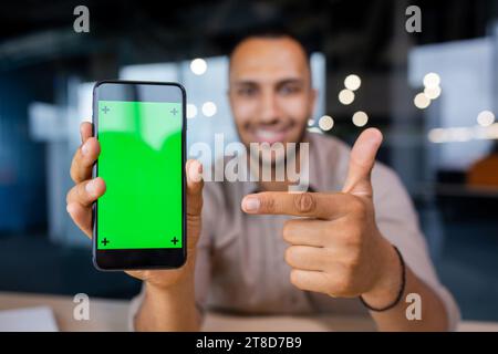Smiling man holding a phone with a green screen in his hands, showing it to the camera and pointing at it with his index finger, looking at the camera. Stock Photo